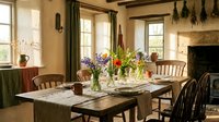 Rustic farmhouse dining room with wooden table, fresh wildflowers, dried herbs hanging from beamed ceiling, and warm natural light.