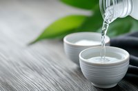 Pouring clear liquid from glass bottle into white ceramic cups on wooden surface with green leaves and spa stones creating zen wellness atmosphere.