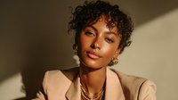 Young woman with curly hair and gold jewelry in warm sunlight wearing beige blazer, showcasing natural makeup and elegant style for fashion or beauty campaigns.