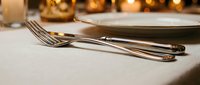 Elegant table setting with polished silverware, white napkin, and warm golden bokeh lights in background, suitable for fine dining, restaurant, catering, and event design projects.