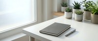 Minimalist white desk workspace with closed notebook, pen and small potted plants by window. Clean modern home office setup for productivity and focus.
