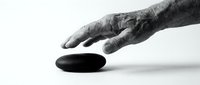 Weathered hand reaching toward smooth black stone on white surface, conveying touch, balance, and minimalist zen concept for meditation and wellness design.