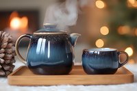Steaming ceramic teapot and cup set in blue glaze finish on wooden tray with cozy bokeh lights and pinecone, creating warm winter atmosphere for tea ceremony.