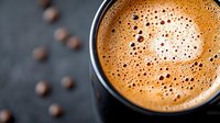 Fresh brewed espresso with golden crema foam in black cup on dark background with scattered coffee beans, macro close-up view of bubbles.