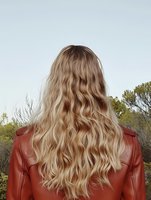 Young blonde woman with long wavy hair wearing red leather jacket viewed from behind against natural outdoor background with trees and sky.