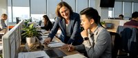 Female manager with brown hair and Caucasian ethnicity mentoring young male employee at computer workstation in modern office environment with colleagues working in background.