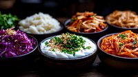 Variety of colorful Korean side dishes banchan in traditional bowls including kimchi, purple cabbage, rice and creamy dip garnished with fresh herbs and sesame seeds.