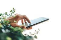 Close-up of Caucasian hand using digital tablet surrounded by green plants, creating natural tech environment for business or lifestyle content.