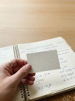 Hand holding blank adhesive note over open spiral notebook with mathematical equations and formulas on wooden desk surface for studying.