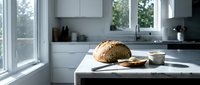 Freshly baked artisan bread loaf with sliced pieces and olive oil on white marble kitchen counter with modern minimalist interior design.