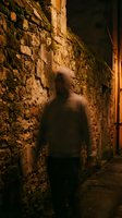 Blurred figure in dark hoodie walking through narrow alley with weathered stone walls at night, creating mysterious and atmospheric urban scene for thriller and suspense projects.