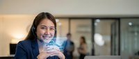 Smiling Asian female customer support agent wearing headset holds glowing 24/7 holographic tablet in modern call center office.