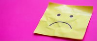 Yellow sticky note with sad face emoticon on bright pink background expressing disappointment and negative emotions for office communication.