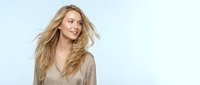 Young blonde woman with flowing hair in beige sweater smiling joyfully against light blue background. Natural beauty and happiness concept for lifestyle content.