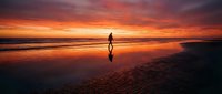 Silhouette of male surfer holding surfboard standing on wet sand beach at dramatic golden sunset with vibrant orange sky reflecting on water.