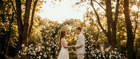 Romantic wedding ceremony with bride and groom exchanging vows under floral arch in golden sunset forest setting with warm natural lighting.