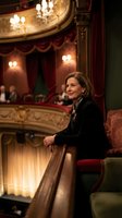 Elegant mature woman in black attire sitting in ornate opera house theater box with red velvet seats and golden chandeliers creating luxurious cultural atmosphere.