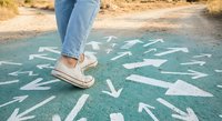 Person in casual sneakers standing on asphalt with white directional arrows pointing in multiple directions, symbolizing choice and decision-making concepts.