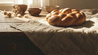 Freshly baked braided challah bread on rustic wooden table with warm sunlight streaming through window creating cozy kitchen atmosphere for bakery marketing.