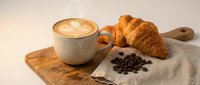 Fresh croissant and latte coffee with latte art on wooden board with coffee beans. Morning breakfast setup for cafe menu and food photography.