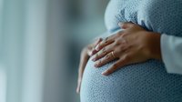 Young pregnant woman in white sweater gently holding her belly, wearing blue polka dot dress. Intimate moment captured in soft natural window light.