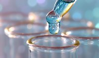 Liquid droplet falling into glass test tube with golden rim against blurred bokeh background, macro photography showing surface tension and fluid dynamics.