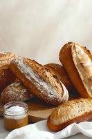 Fresh crusty artisan bread loaves with crispy golden crust and jar of honey on wooden board against light background, rustic food photography styling.