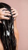 Woman washing long dark hair with shampoo foam in shower against pink background. Hair care routine and personal hygiene concept for beauty products.