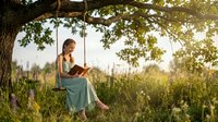Young woman in turquoise dress reading book on tree swing in sunny meadow during golden hour. Peaceful outdoor leisure and literature concept.