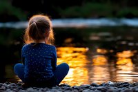 Young girl in blue sweater sitting alone on rocky riverbank at sunset, watching golden reflections on water surface. Peaceful contemplative childhood moment.