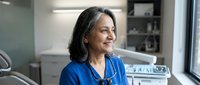 Middle-aged woman with dark hair and South Asian ethnicity wearing blue medical scrubs and stethoscope in modern dental or medical clinic office setting, healthcare professional portrait.