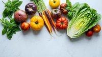 Fresh organic vegetables and herbs arranged on white marble surface including tomatoes, carrots, onions, peppers, leafy greens and basil for healthy cooking.