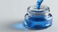Blue liquid droplet falling into clear glass jar with vibrant blue solution, showcasing fluid dynamics in cosmetic or laboratory setting.