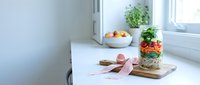 Layered succulent garden in glass jar with pink ribbon on wooden board, fresh oranges in bowl, potted plant by bright window in minimalist kitchen or office workspace.