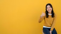 Young woman with long dark hair and East Asian ethnicity wearing mustard yellow sweater giving thumbs up gesture against bright yellow background. Suitable for positive messaging, success concepts, and lifestyle marketing campaigns.