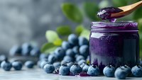 Fresh blueberry smoothie in glass jar with wooden spoon and scattered berries on light surface for healthy breakfast recipes and nutrition content.
