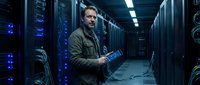 Middle-aged Caucasian male IT technician working with laptop in modern server room data center with blue LED lighting and network equipment racks.