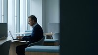 Mature male professional of European descent working on laptop at modern office desk by window, business environment.
