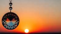 Crystal pendant with geometric facets hanging against golden sunset sky creating spiritual meditation atmosphere for wellness practices.