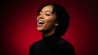 Young African American woman laughing joyfully with eyes closed wearing black turtleneck against vibrant red gradient background expressing happiness and positivity.