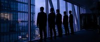 Business team silhouettes standing by office windows overlooking city skyline at dusk. Corporate professionals in suits discussing strategy and planning.