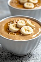 Creamy peanut butter banana smoothie bowl topped with fresh banana slices and oats in white ceramic bowl. Close up view with selective focus.