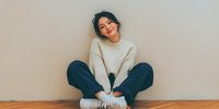 Young woman with dark hair sitting cross-legged on wooden floor wearing cozy cream sweater and dark pants, smiling with eyes closed against neutral wall.