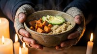 Hands in knitted gloves holding wooden bowl with quinoa, roasted sweet potato and cucumber slices, surrounded by lit candles for wellness and mindful eating content.