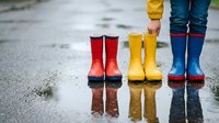 Young children wearing red, yellow, and blue rain boots standing in puddles on wet pavement, ideal for seasonal weather and childhood themes.