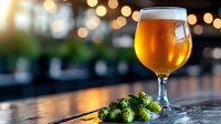 Glass of cold craft beer with fresh green hops on wooden bar counter against blurred bokeh lights background. Golden ale with foam head and bubbles.