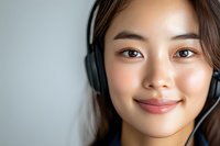 Young Asian woman wearing headphones smiling at camera with natural makeup and glowing skin against light background, expressing joy and confidence.
