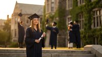 Young blonde woman graduate in cap and gown holding diploma at university campus during golden hour sunset with classmates celebrating in background.