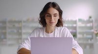 Young Caucasian woman in glasses and white sweater working on laptop at home office desk, smiling while looking at computer screen against blurred background.