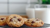 Fresh baked chocolate chip cookies cooling on wire rack with glass of milk in background, warm golden brown treats with melting chocolate morsels.